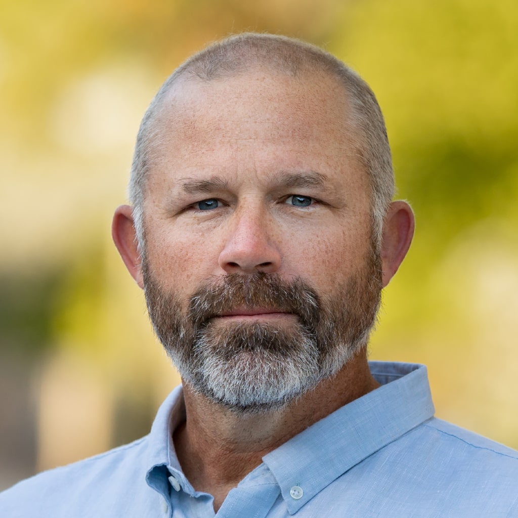 A man with a beard and mustache, wearing a light blue collared shirt, stands in front of a blurred background with shades of green and yellow, reminiscent of the welcoming ambiance found in dog daycare facilities.