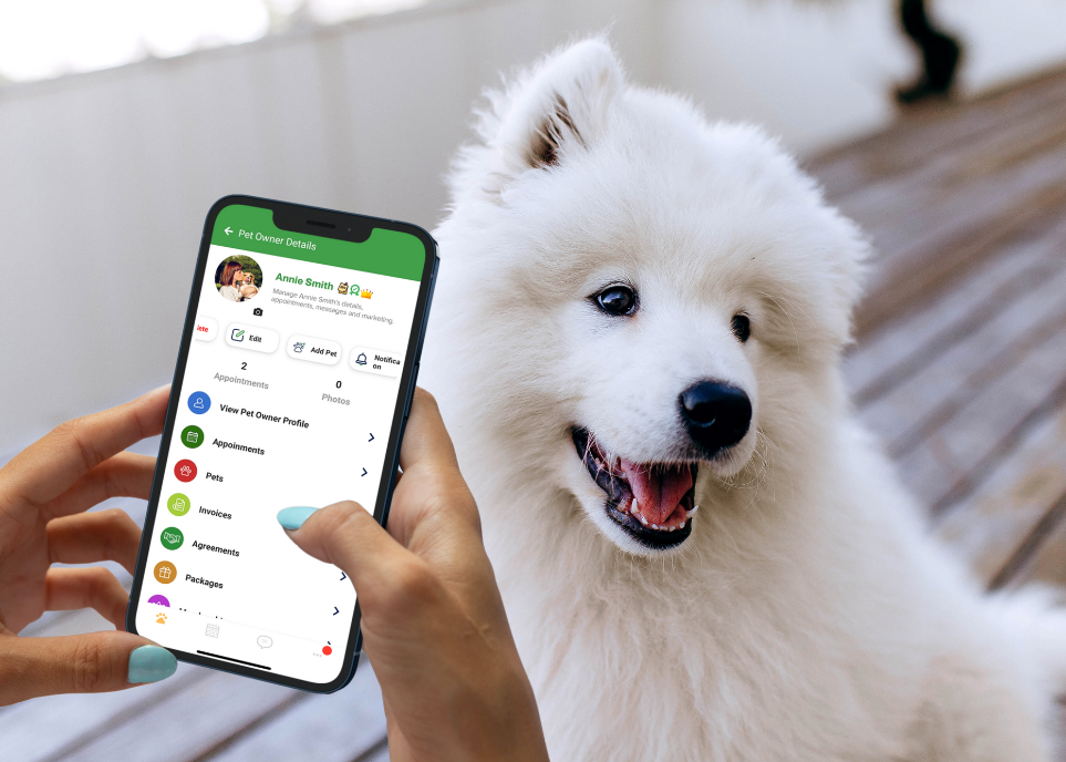 A person holding a smartphone with the RunLoyal pet care app open, displaying pet owner details. At home on a wooden floor, a fluffy white dog sits contentedly next to the person.