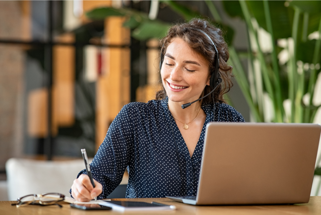 A woman wearing a headset smiles while taking notes in front of a laptop, embodying the perfect blend of professionalism and comfort at home. Glasses rest on the table, with lush green plants creating a serene backdrop, enhancing her focus and productivity.